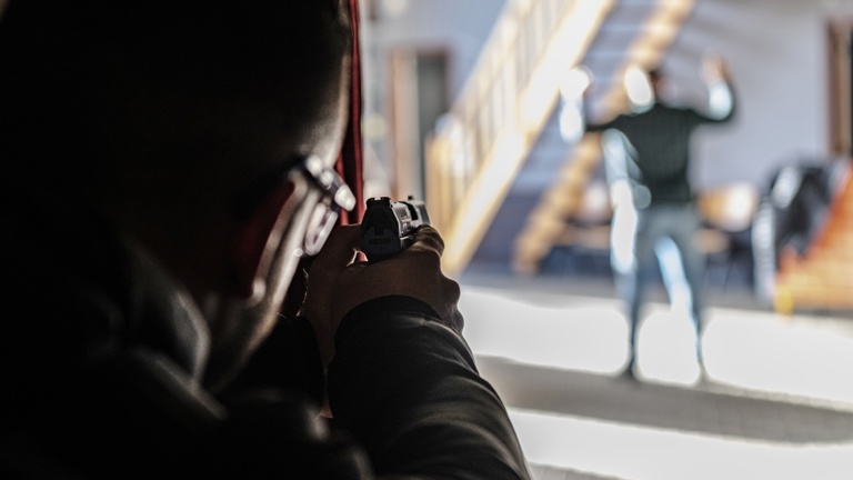 Politieacademie studierichting Bijzondere opsporing -Silhouet van persoon die wapen richt op persoon op achtergrond met handen in de lucht