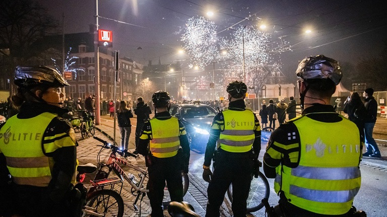 Vier agenten bekijken op een afstand een groep mensen met vuurwerk op de achtergrond tijdens de jaarwisseling