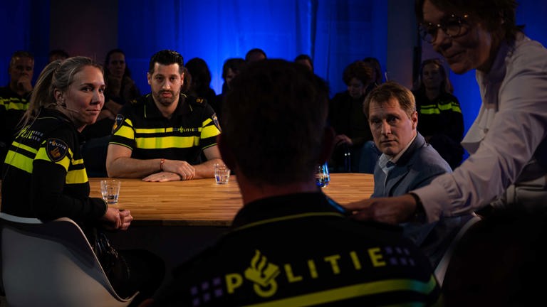 Politiemensen aan tafel in een debat. De leider van het debat geeft een politieman de microfoon.