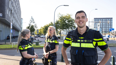 Politiestudent lacht met handen in zij naar de camera, op de achtergrond praten twee politiestudenten met elkaar