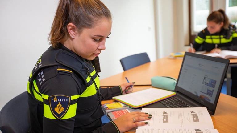 Een student in politie-uniform zit achter de laptop en kijkt in een studieboek