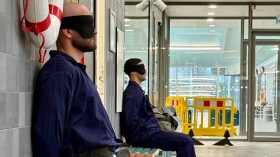 Two students dressed in blue overalls and blindfolded on a bench.